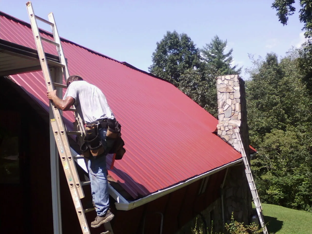 Roofer on a ladder beside a red metal roof and stone chimney — Soffit & Fascia Repair by Roofing Brothers in Rancho Cucamonga